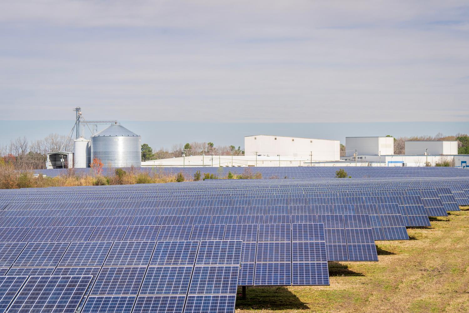 Parque solar fotovoltaico con naves industriales al fondo, representando la estructura de un PPA Off-site. Parque solar fotovoltaico con naves industriales al fondo, representando la estructura de un PPA Off-site.