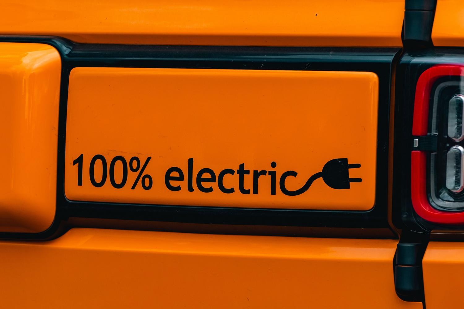 Primer plano del emblema '100% electric' en la parte trasera de un vehículo eléctrico de color naranja vibrante.