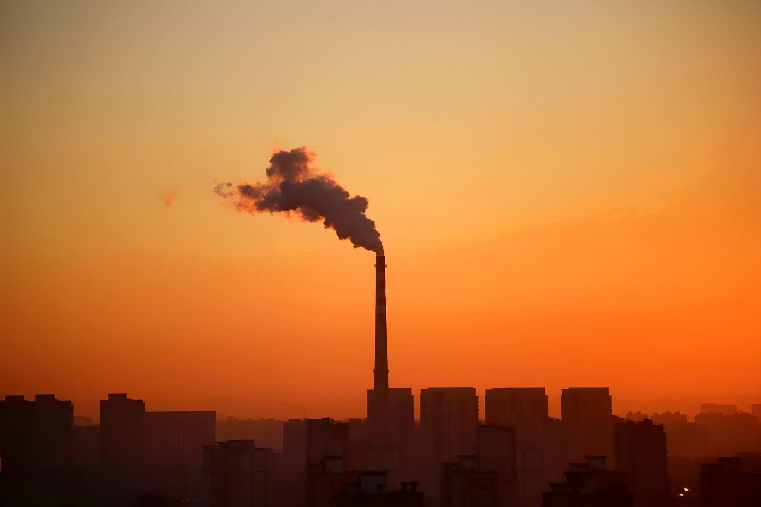 Chimenea industrial emitiendo una densa columna de humo sobre el perfil urbano, representando la emisión de contaminantes primarios y polución industrial.