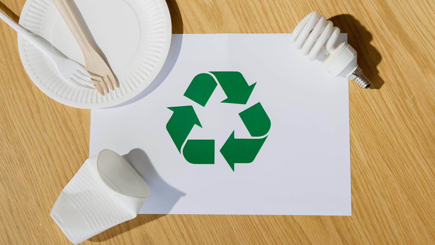 Conjunto de utensilios ecológicos, bombilla y símbolo de reciclaje sobre papel, representando la sostenibilidad en el diseño de productos.