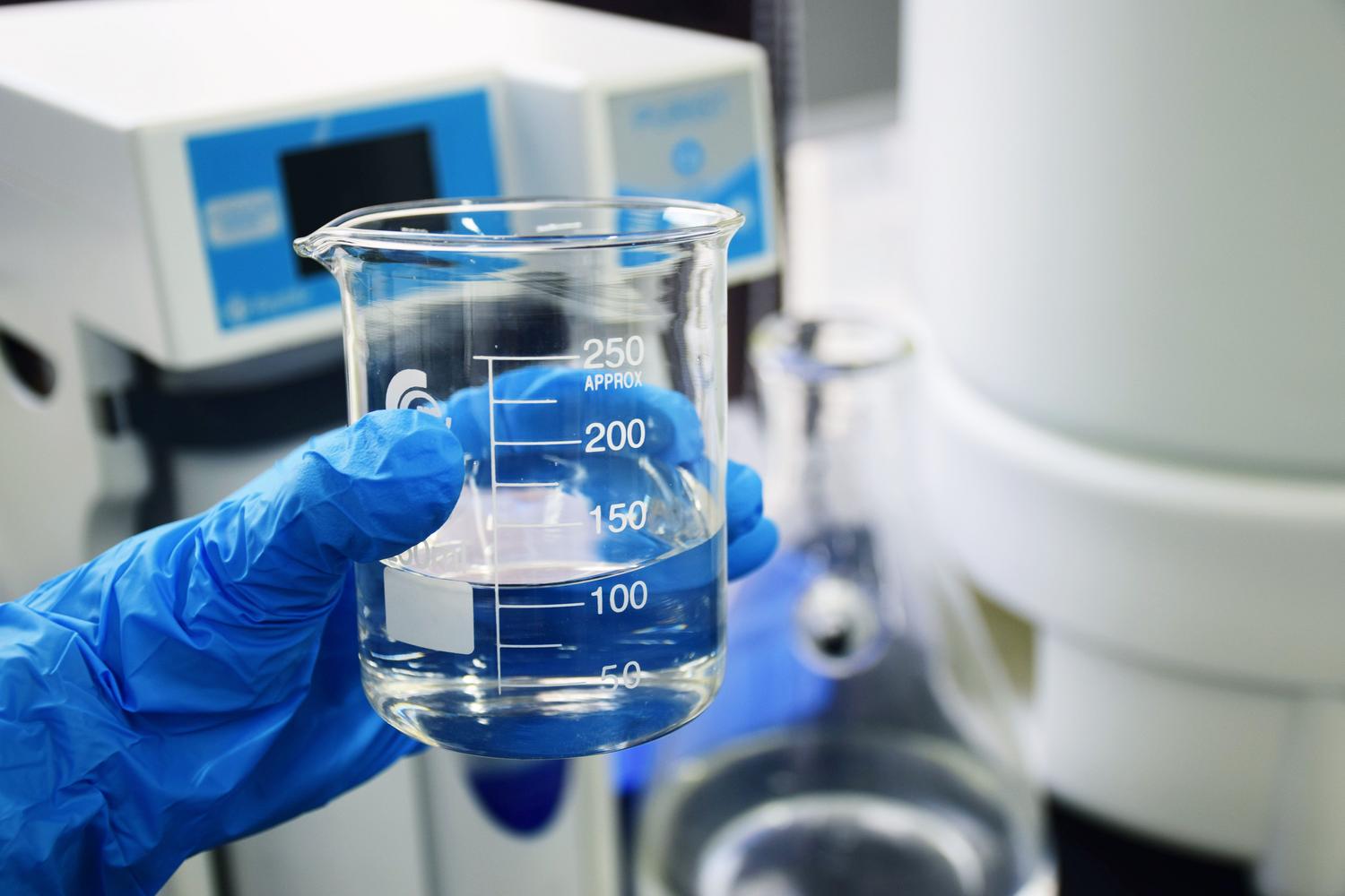 Mano con guantes de protección sosteniendo un vaso de precipitados con una muestra de agua en un laboratorio.