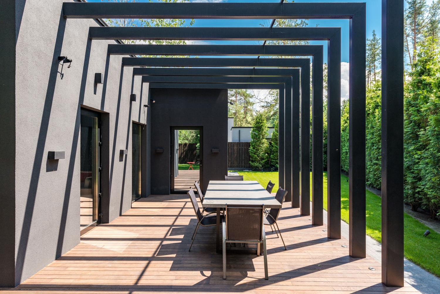Pérgola moderna de diseño geométrico en color oscuro sobre suelo de madera, creando zonas de sombra en una terraza
